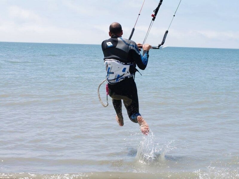 Homme en combinaison noire lançant un saut en kitesurf depuis le bord de l'eau sur une plage ensoleillée.