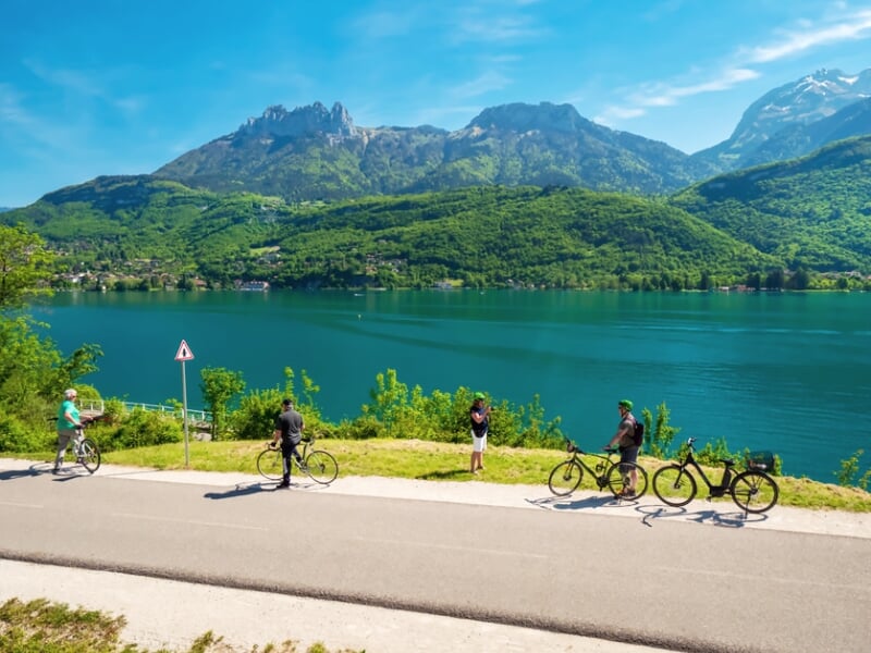 Quatre personnes avec vélos électriques sur une route au bord d'un lac entouré de montagnes verdoyantes sous un ciel bleu.