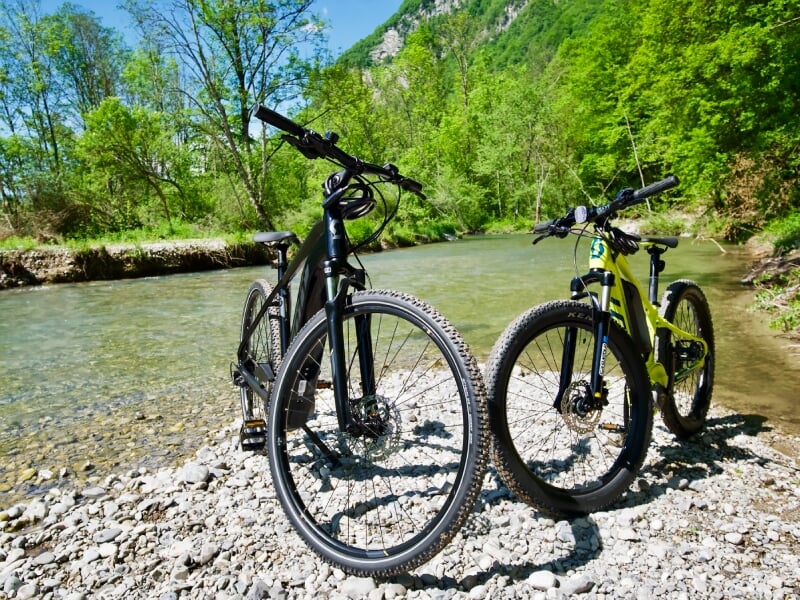 Deux vélos électriques posés sur des cailloux au bord d'une rivière claire avec une forêt dense en arrière-plan.