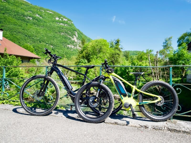 Deux vélos électriques stationnés sur une route près d'une barrière verte avec des collines et des arbres en arrière-plan.