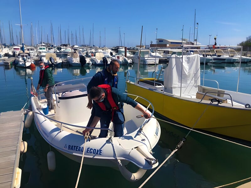 Deux personnes en gilets de sauvetage sur un petit bateau gonflable blanc dans un port avec de nombreux voiliers.