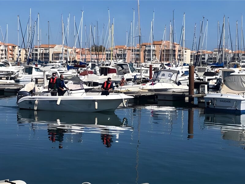 Deux personnes en gilets rouges naviguent sur un petit bateau blanc dans un port calme avec des bateaux amarrés.