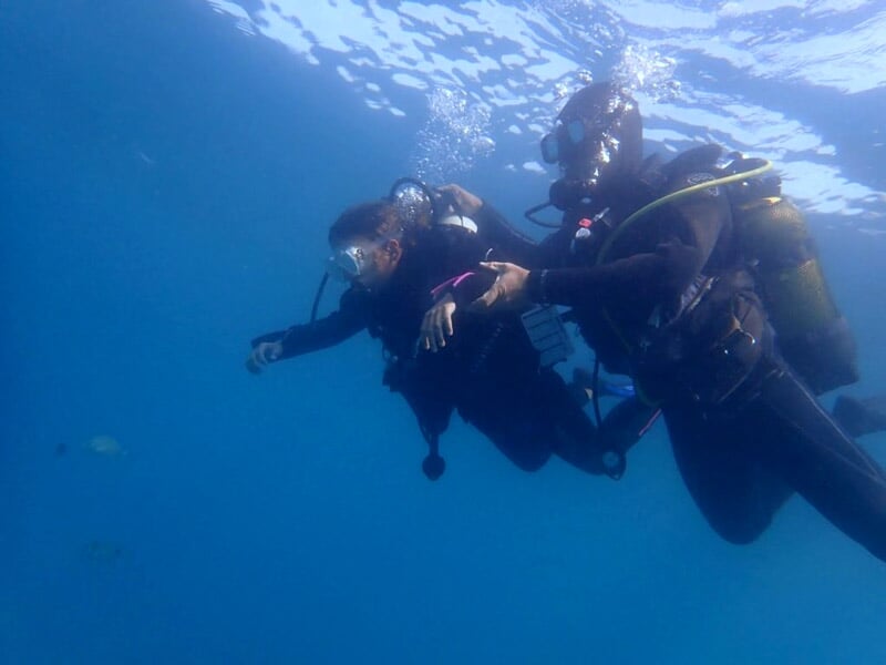 Un instructeur guide une plongeuse sous l’eau, les deux portant masque, détendeur et gilet stabilisateur, dans une mer calme.