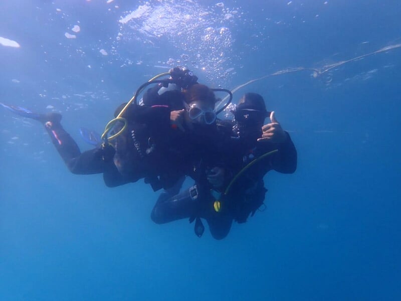 Deux plongeurs en combinaison et équipement de plongée se tiennent sous l’eau, entourés de bulles, l’un d’eux faisant un signe de la main.