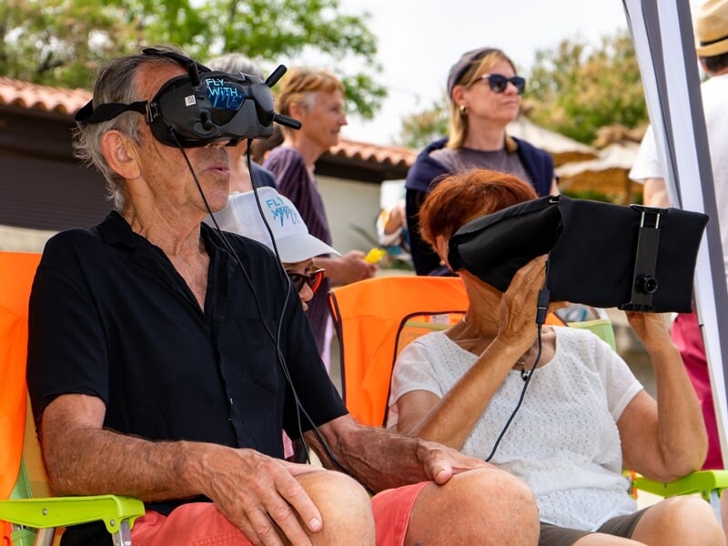 Deux personnes âgées assises portant des casques de réalité virtuelle lors d'une activité en extérieur sous un ciel clair.
