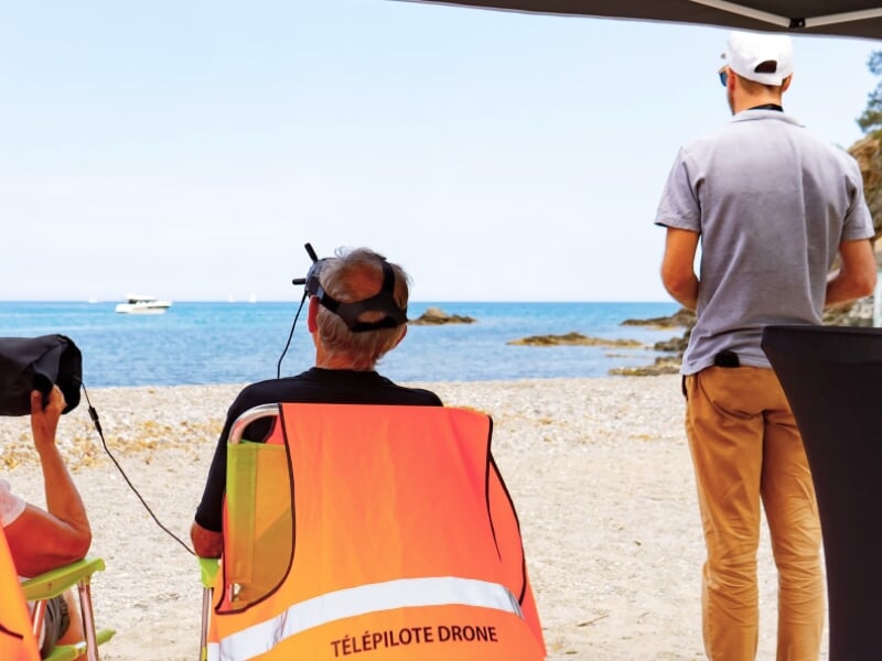 Deux hommes sur une plage, l'un assis avec un casque de réalité virtuelle et l'autre debout, près de la mer calme.