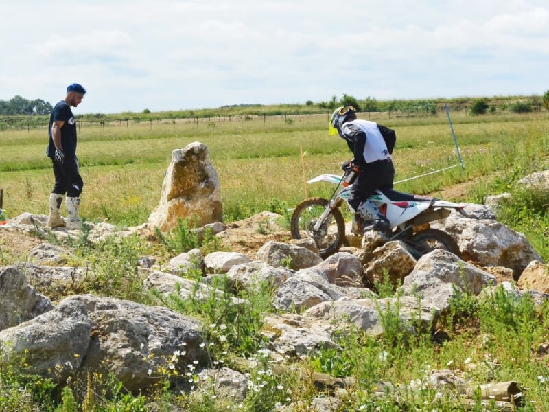 Un pilote en tenue complète sur une moto tout-terrain franchit des rochers sous la supervision d'un moniteur.