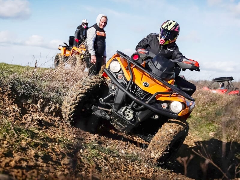 Groupe de personnes en quad orange évoluant sur un terrain accidenté avec boue et végétation.