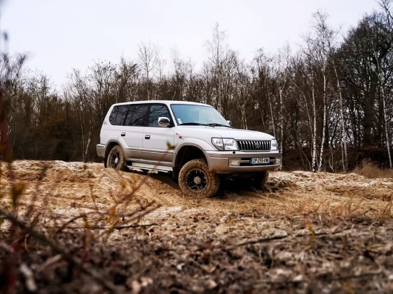 Véhicule 4x4 blanc évoluant sur un terrain boueux et accidenté avec des arbres en arrière-plan.