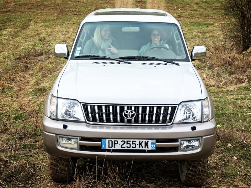 Vue avant d'un 4x4 blanc avec deux personnes à bord sur un chemin de terre en zone rurale.