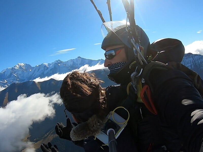 Deux personnes en parachute lors d'un saut en tandem au-dessus des montagnes enneigées sous un ciel bleu clair.