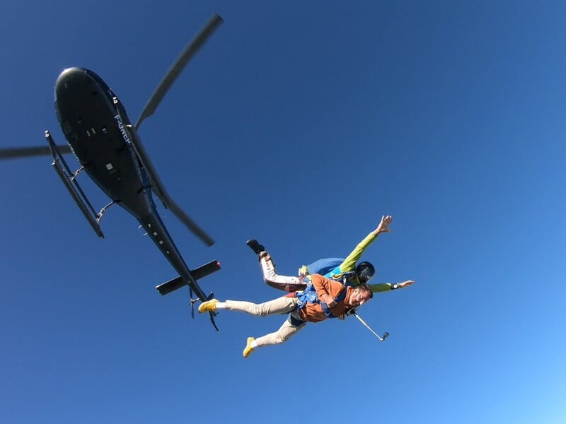 Deux personnes effectuent un saut en parachute en tandem depuis un hélicoptère dans un ciel bleu clair sans nuages.