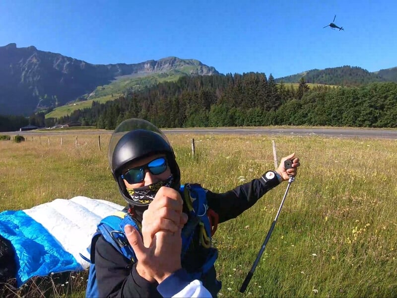 Parachutiste au sol en pleine nature, casque et lunettes, tenant une perche selfie après un saut en parachute réussi.