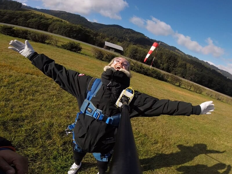 Personne souriante en combinaison de parachutisme avec harnais bleu, bras ouverts sur un champ vert sous un ciel bleu.