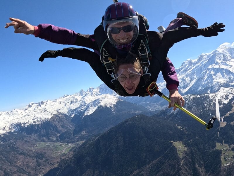 Deux personnes en parachute en tandem survolent les montagnes enneigées du Massif du Mont-Blanc sous un ciel bleu clair.