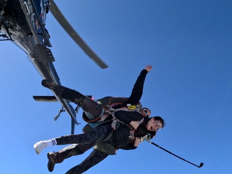 Personne en parachute en tandem sautant d'un hélicoptère avec ciel bleu clair en arrière-plan, expression de joie visible.