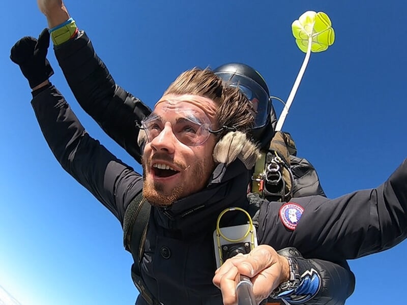 Tandem de parachutistes en chute libre, le passager souriant avec un casque, lunettes et gilet, ciel bleu clair.