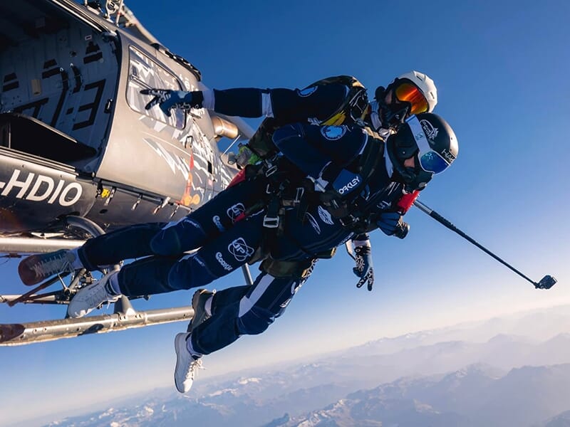 Deux parachutistes en tandem sautent d'un hélicoptère au-dessus des montagnes sous un ciel bleu clair.