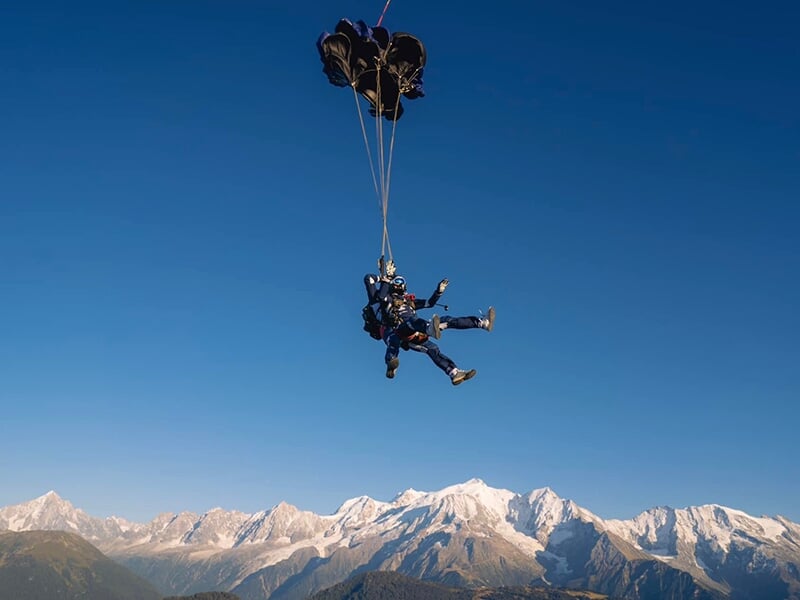 Parachutistes descendant sous un parachute noir, montagnes enneigées majestueuses en arrière-plan.