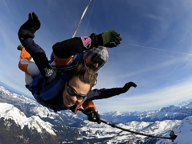 Parachutistes volant côte à côte au-dessus des nuages, montagnes enneigées visibles à l'horizon.