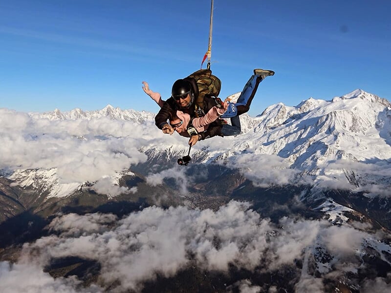 Parachutistes volant côte à côte au-dessus des nuages, montagnes enneigées visibles à l'horizon.