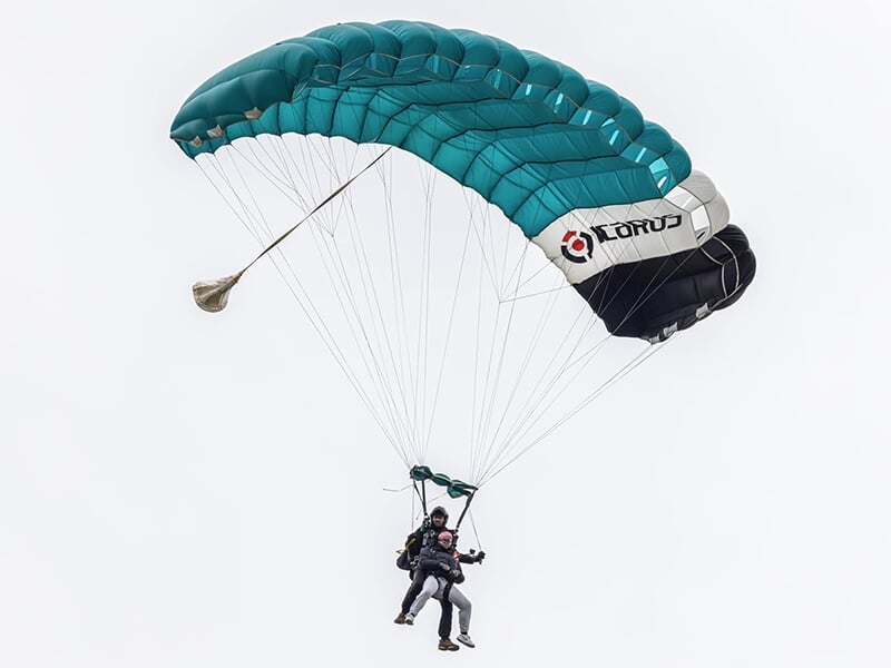 Tandem de parachutistes descendant sous un grand parachute vert et noir, avec un ciel gris clair en arrière-plan.