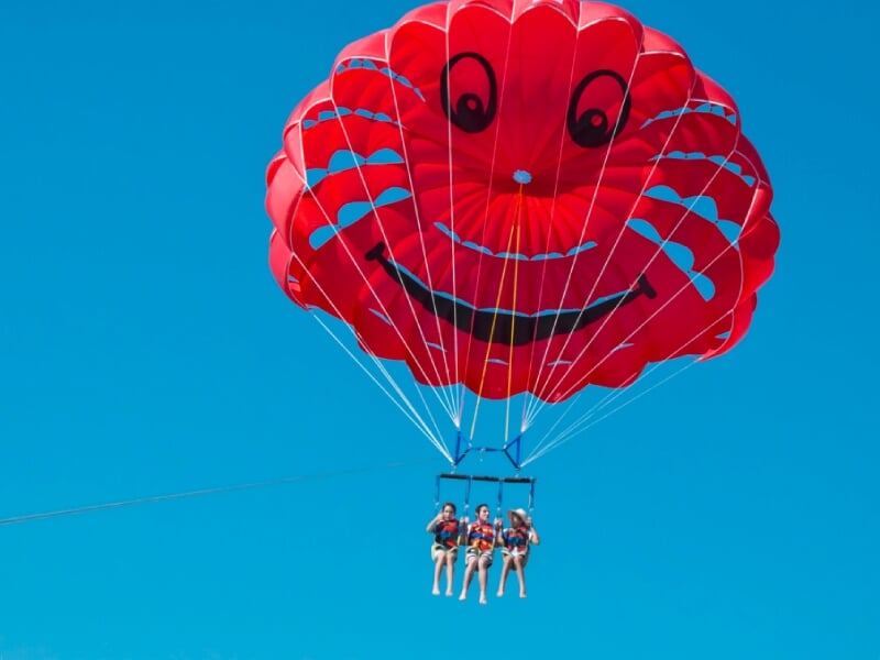 Trois personnes en parachute ascensionnel sous un parachute rouge souriant au-dessus de la mer bleue claire.