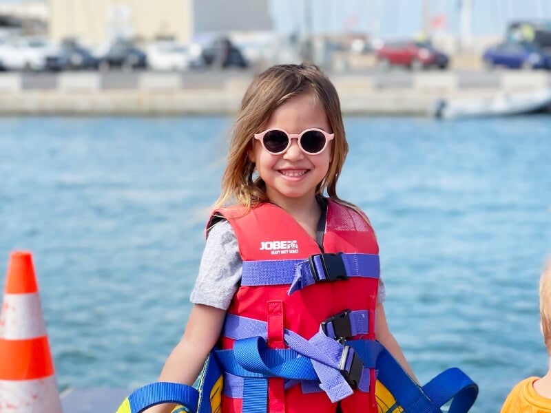 Jeune fille souriante portant un gilet de sauvetage rouge et des lunettes de soleil, debout sur un quai au bord de l'eau.