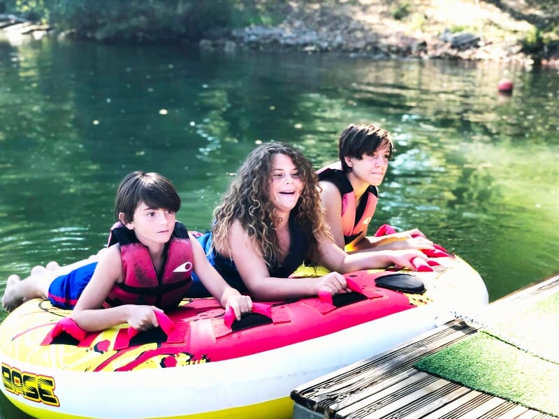 Trois enfants portant des gilets de sauvetage sur une bouée tractée rouge et jaune au bord de l'eau.