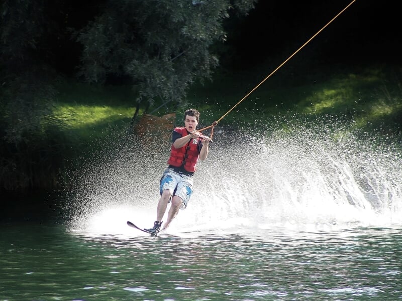 Jeune garçon en short bleu pratiquant le ski nautique sur un lac entouré d'arbres verts.