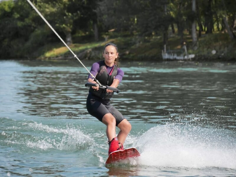 Femme en combinaison de plongée faisant du wakeboard sur un lac calme avec des arbres en arrière-plan.