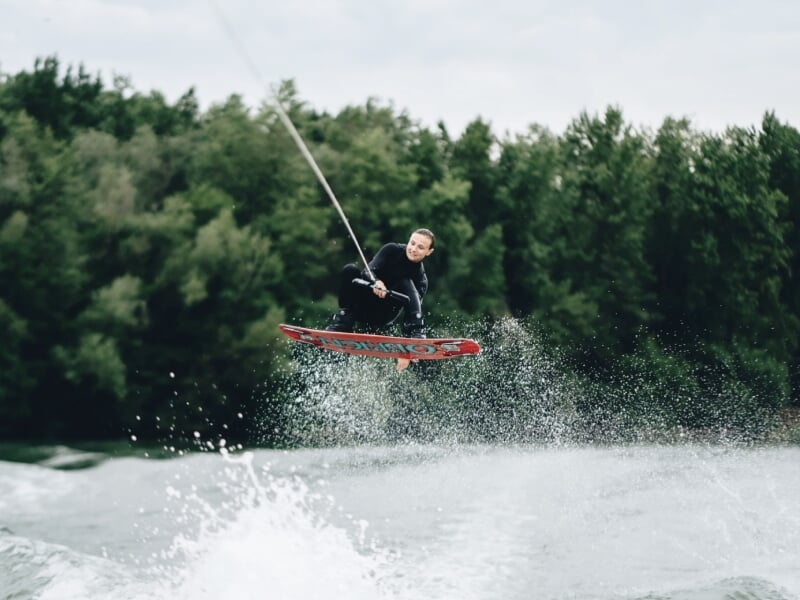 Homme en combinaison noire effectuant un saut spectaculaire en wakeboard sur un lac boisé.