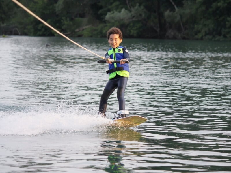 Jeune garçon souriant en gilet de sauvetage vert et bleu faisant du wakeboard sur l'eau.