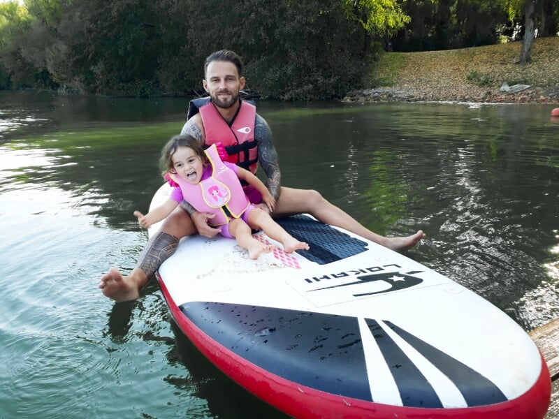 Homme barbu assis sur un paddle avec une petite fille en gilet de sauvetage rose sur un lac.