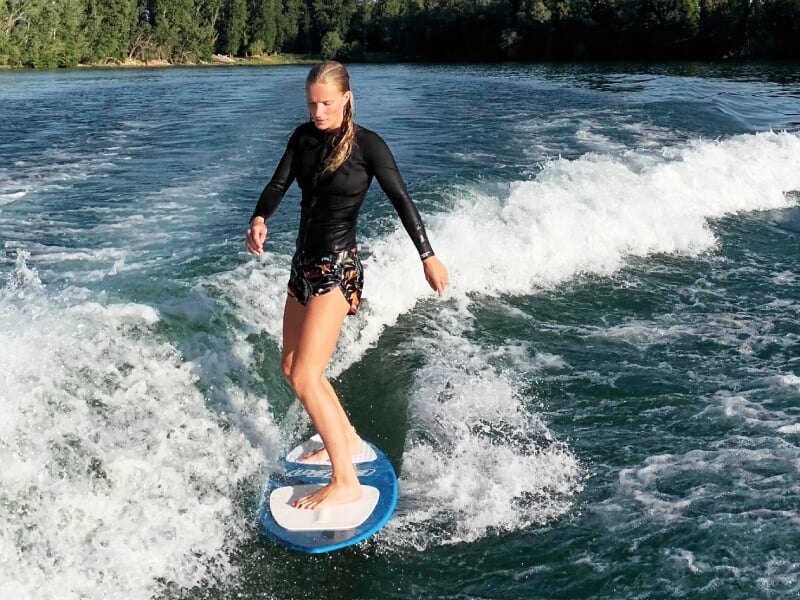 Femme en maillot noir surfant sur une planche de wakesurf sur une vague formée par un bateau.