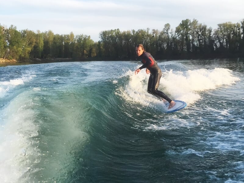 Personne en combinaison noire surfant sur une planche de wakesurf sur une grande vague bleue.