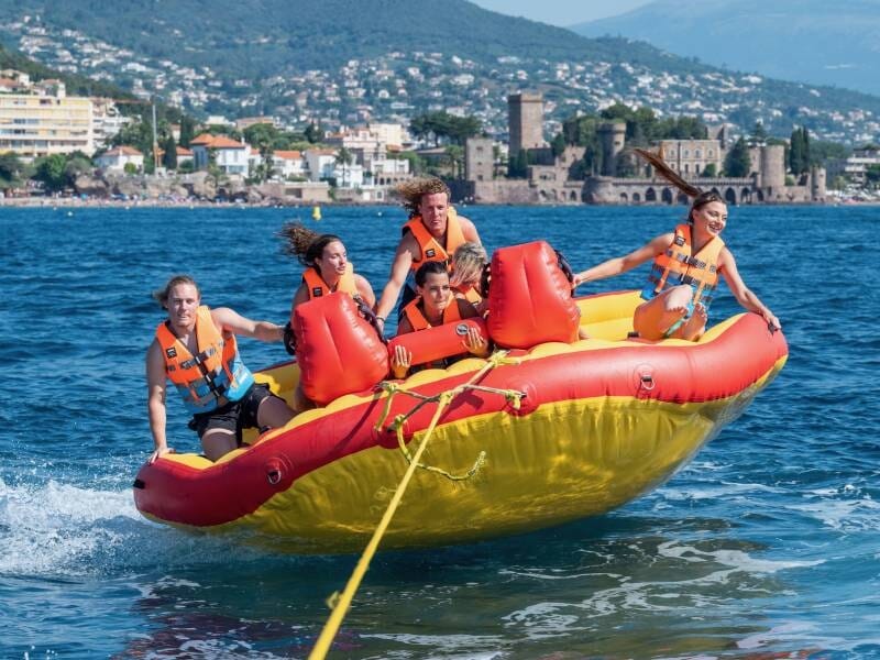 Groupe de cinq personnes en gilets de sauvetage sur une bouée tractée rouge et jaune, avec vue sur la côte et les montagnes, à Mandelieu-la-Napoule.
