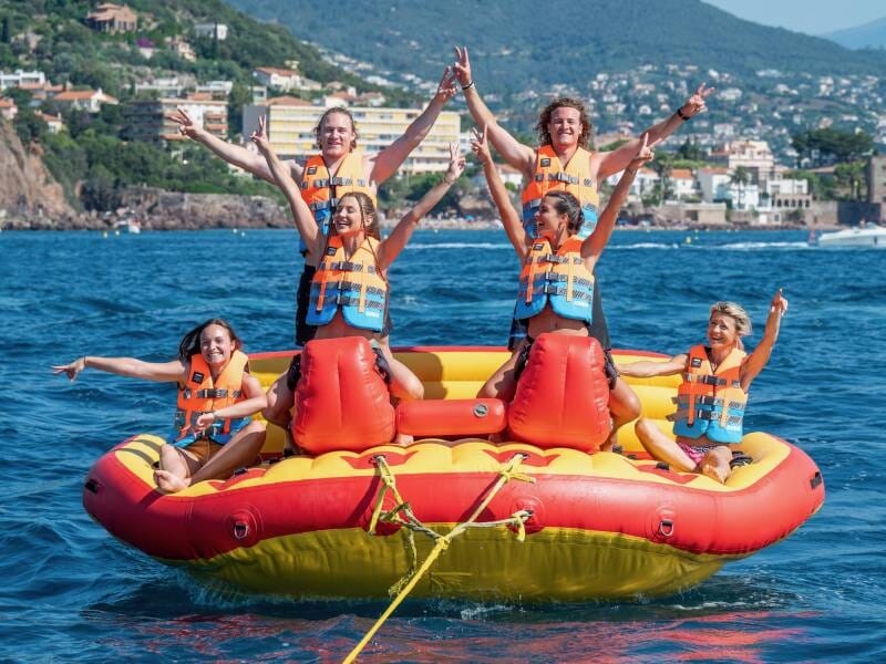 Six personnes souriantes en gilets de sauvetage sur une bouée tractée rouge et jaune, bras levés, en mer devant Mandelieu-la-Napoule.
