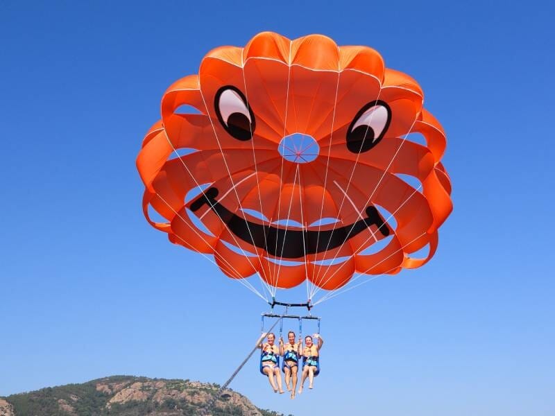 Trois personnes en parachute ascensionnel attachées à un parachute orange souriant au-dessus de la mer bleue.