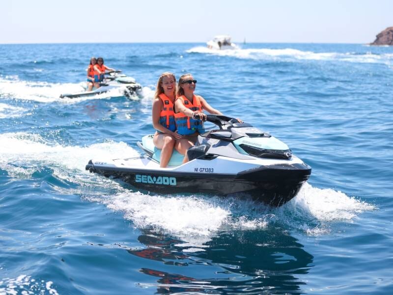 Deux femmes souriantes en gilets orange pilotent un jetski blanc sur une mer bleue avec une autre embarcation en arrière-plan.