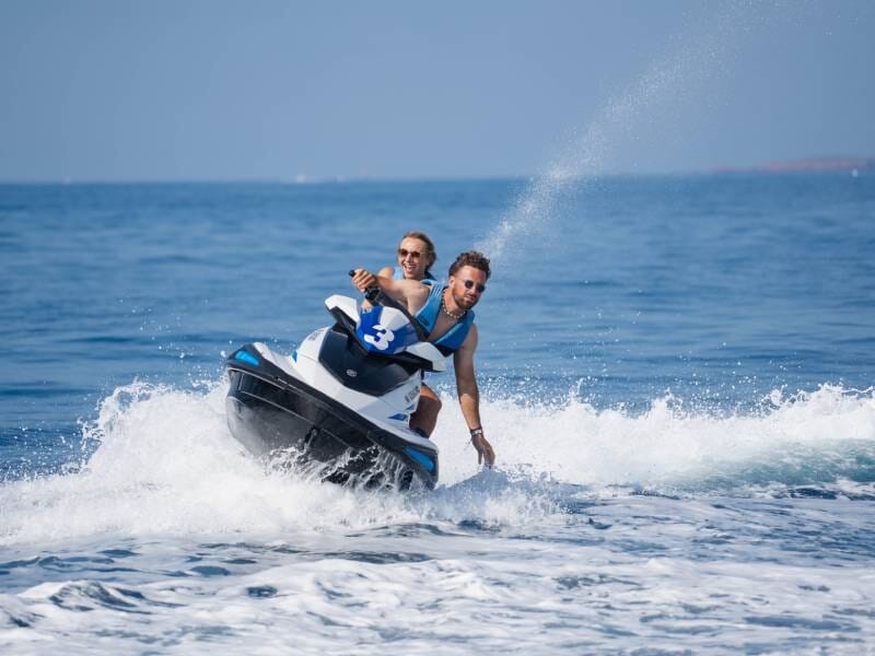 Un homme et une femme en gilets bleus font une virage serré en jetski sur une mer agitée sous un ciel clair.