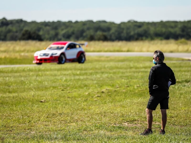Un homme debout sur l'herbe regarde une voiture de course rouge et blanche sur un circuit en arrière-plan.