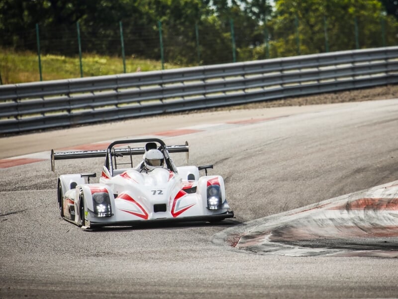 Voiture de course blanche avec casque de pilote sur un virage serré d'un circuit automobile entouré de barrières de sécurité.