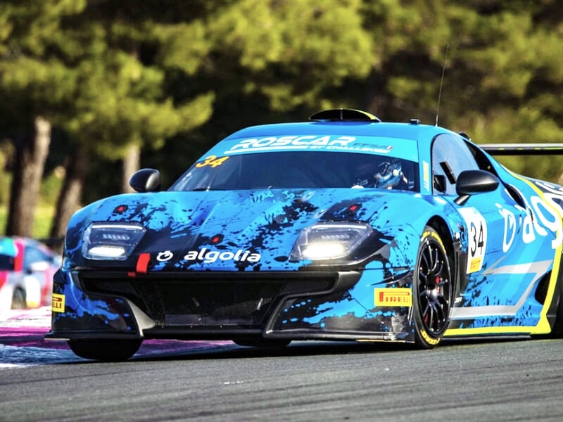 Voiture de course bleue avec motifs noirs et jaunes roulant sur un circuit, entourée d'arbres et d'autres voitures en arrière-plan.