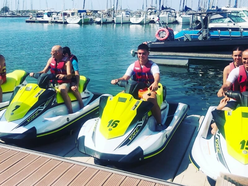 Trois personnes sur des jetskis jaunes et blancs amarrés au quai dans un port avec des bateaux en arrière-plan.