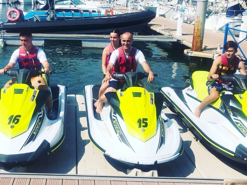 Quatre personnes sur des jetskis jaunes et blancs alignés au quai dans un port avec un bateau noir à l'arrière.