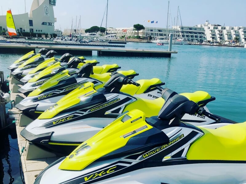 Rangée de jetskis jaunes et blancs stationnés au port avec des voiliers et des bâtiments en arrière-plan.