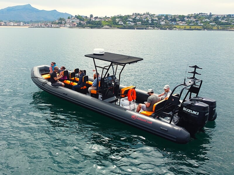 Un bateau pneumatique noir avec plusieurs passagers naviguant sur une mer calme près d'une côte urbaine.