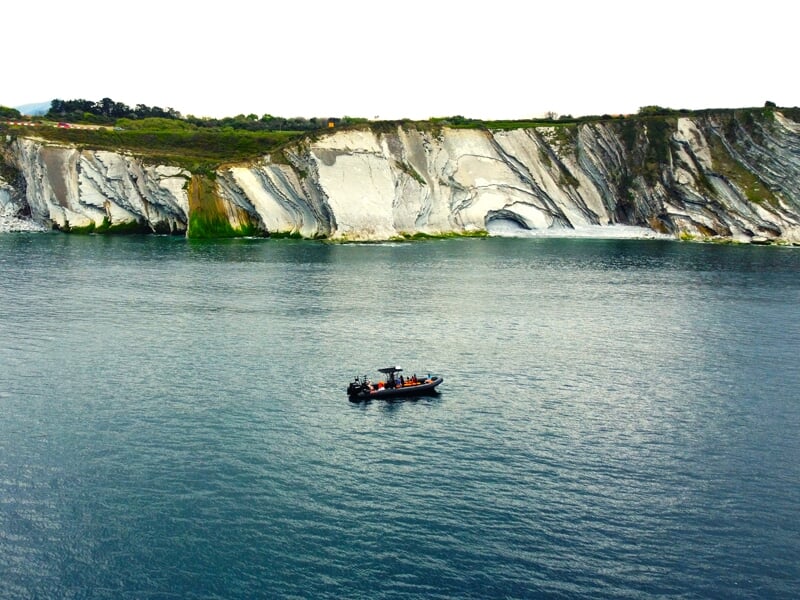 Un petit bateau sur une mer calme avec en arrière-plan une falaise rocheuse blanche et des collines verdoyantes.