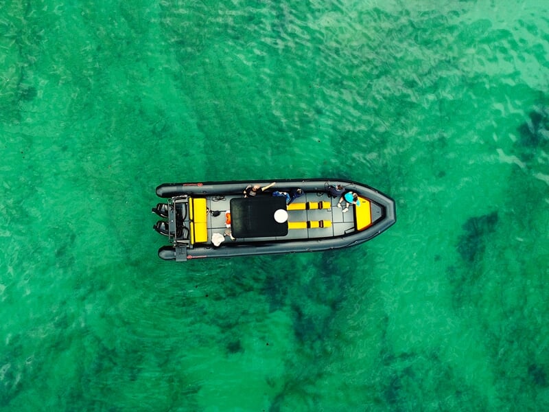 Vue aérienne d'un bateau pneumatique noir avec des sièges jaunes sur une eau turquoise claire.